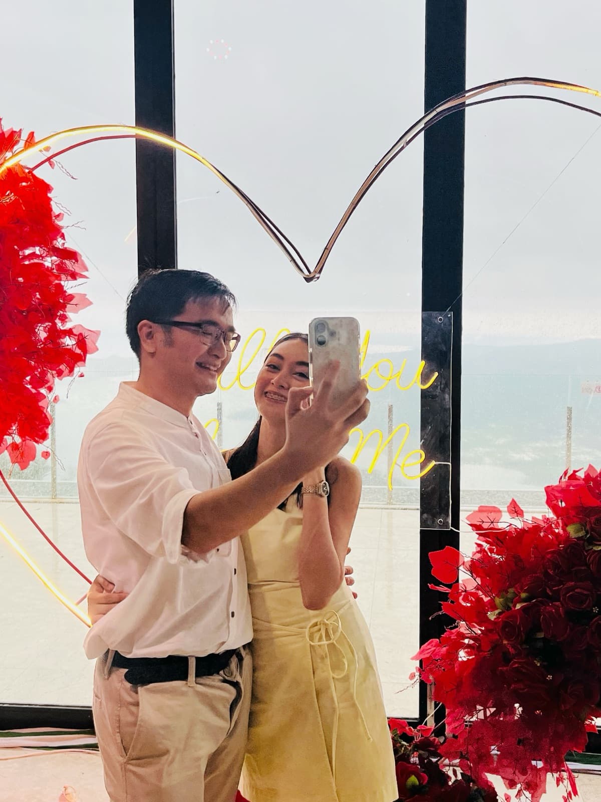 Justin and Lovely taking a selfie together in front of a lit heart-shaped proposal sign, surrounded by red roses
