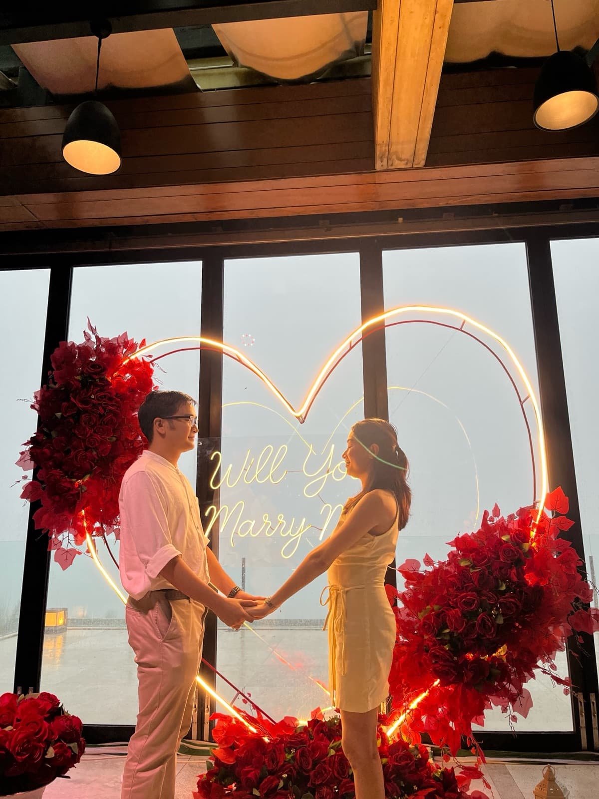 Justin and Lovely holding hands and smiling at each other in front of a lit "Will You Marry Me" heart sign with red rose arrangements