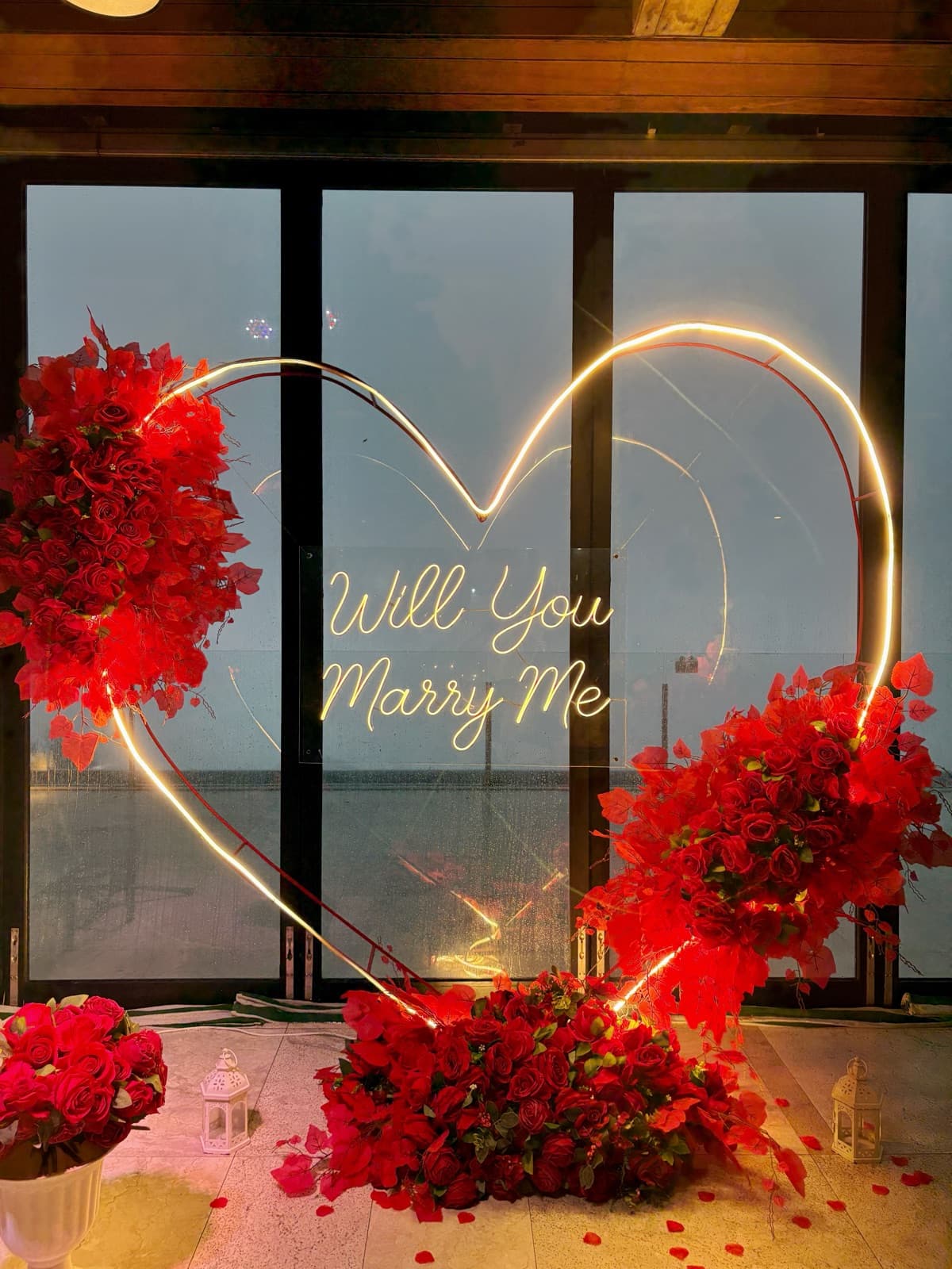 Close-up of the lit heart-shaped "Will You Marry Me" neon sign framed by red rose arrangements and string petals on the floor