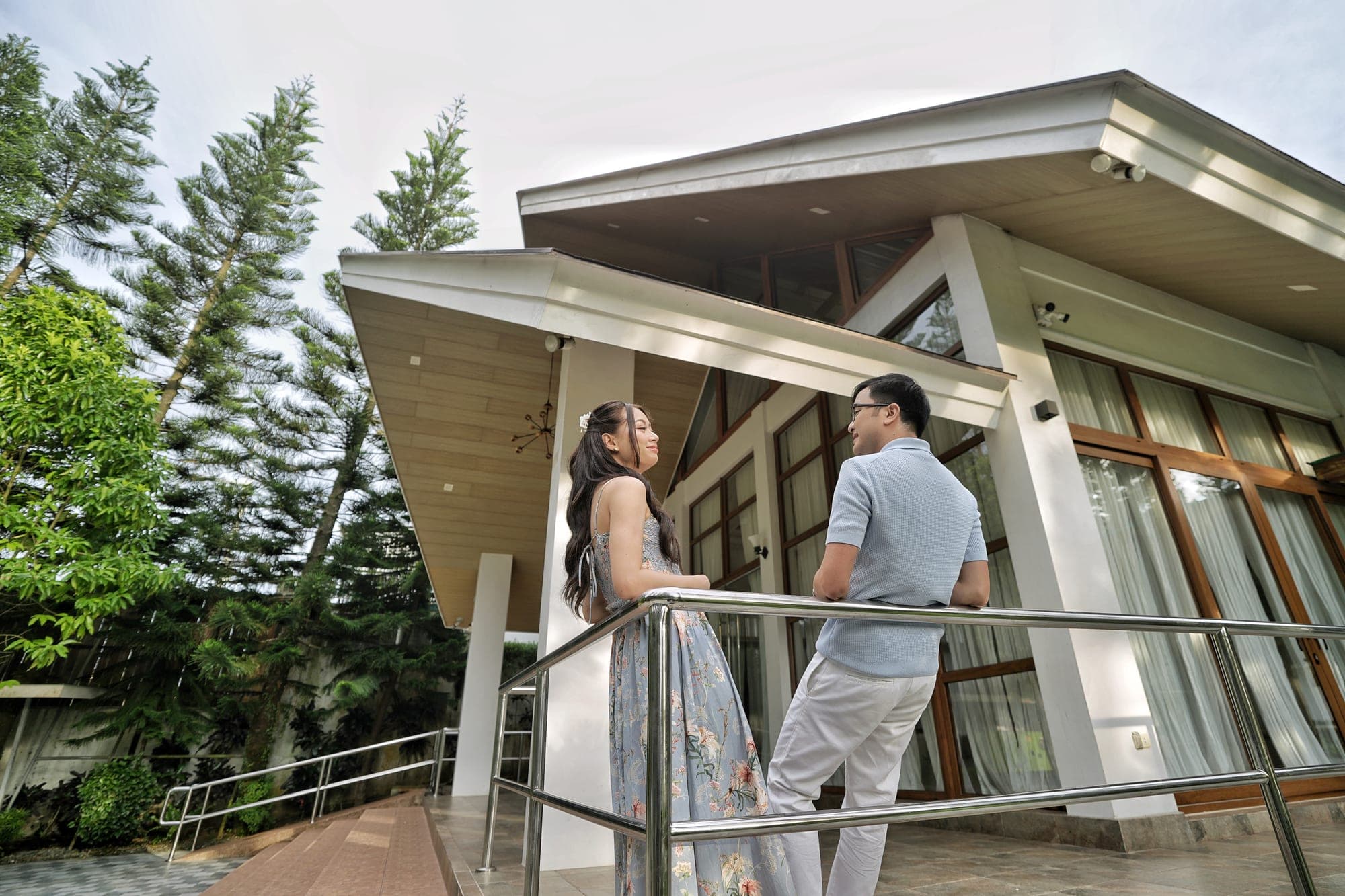 Justin, in a light blue polo, and Lovely, in a floral dress, standing on a balcony rail in front of an angular timber-roofed pavilion