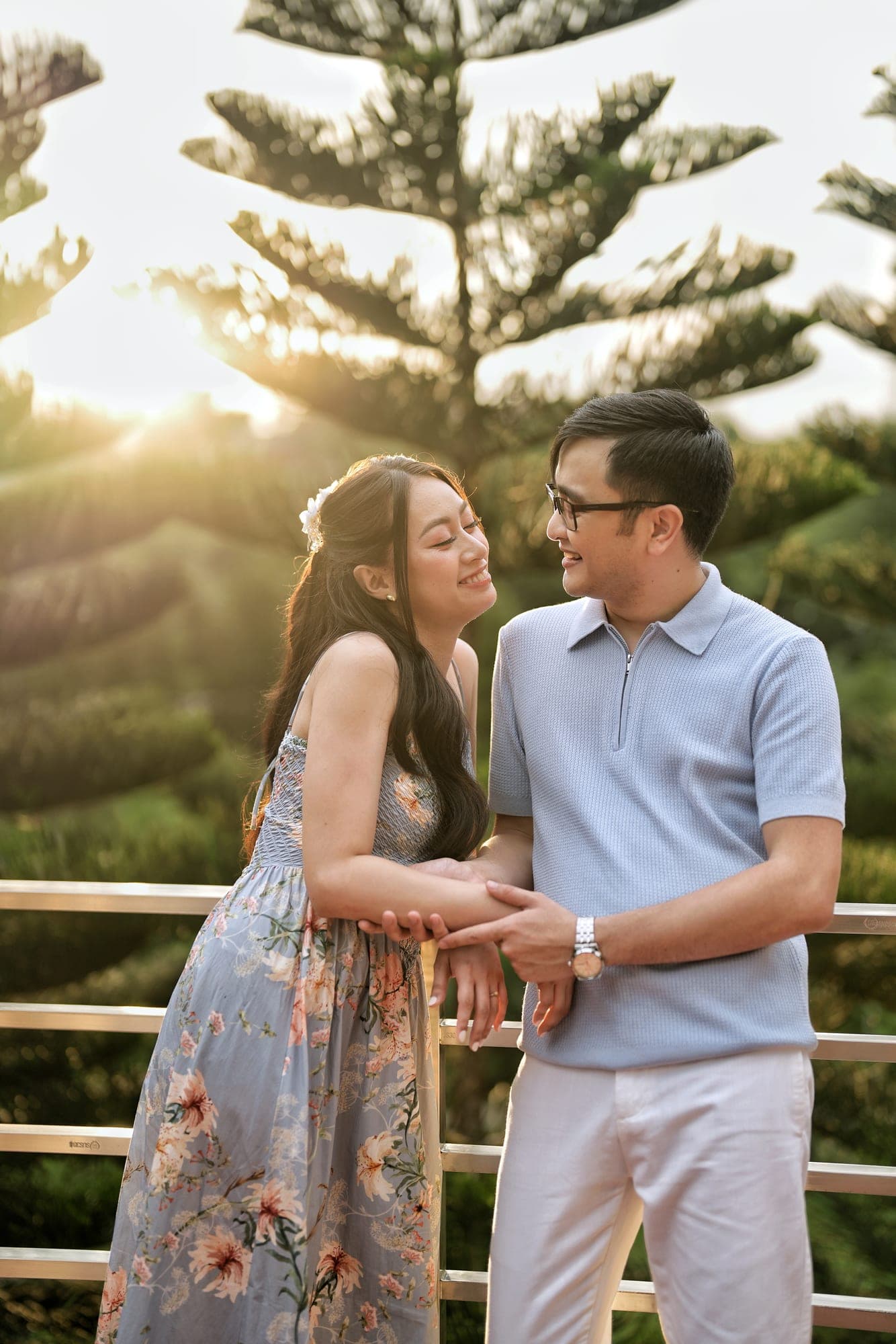 Justin and Lovely smiling at each other at golden hour, leaning on a railing in front of tall pine trees, Lovely in a floral dress