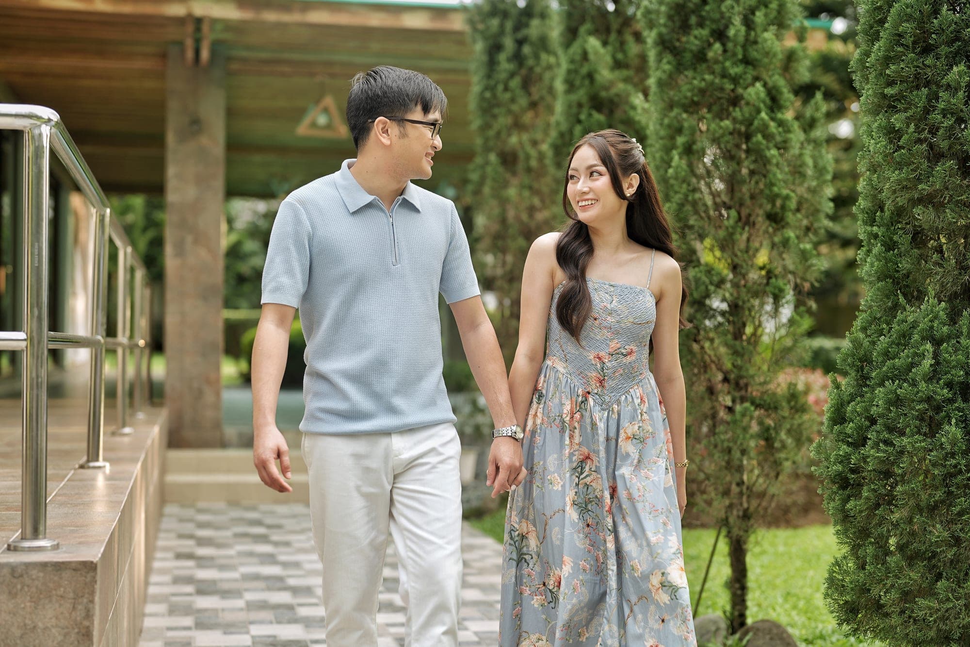 Justin and Lovely holding hands and smiling at each other while walking along a garden pathway lined with topiary trees