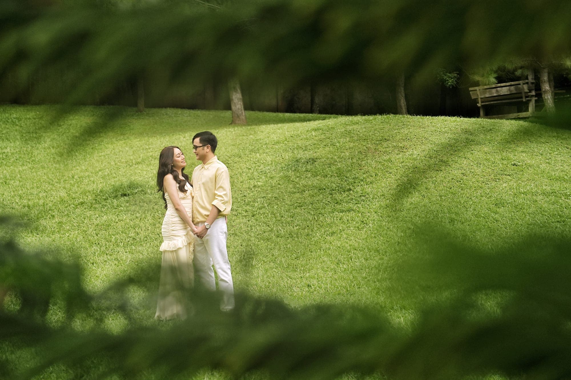 Justin and Lovely, in matching pale yellow outfits, standing together on a grassy hill framed by overhanging pine branches