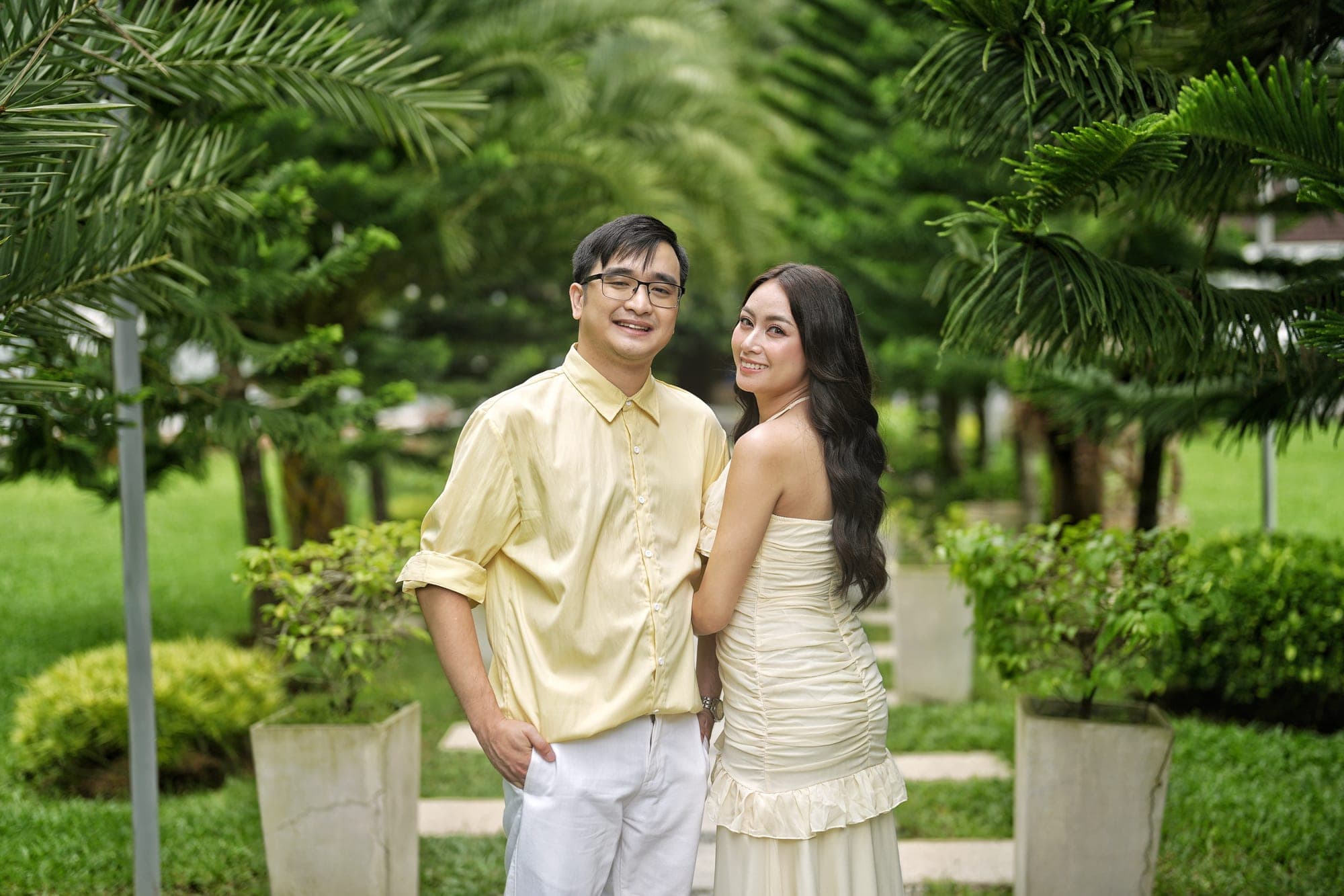 Justin and Lovely, both in pale yellow, standing arm in arm on a garden path lined with potted palms, Lovely glancing back at the camera