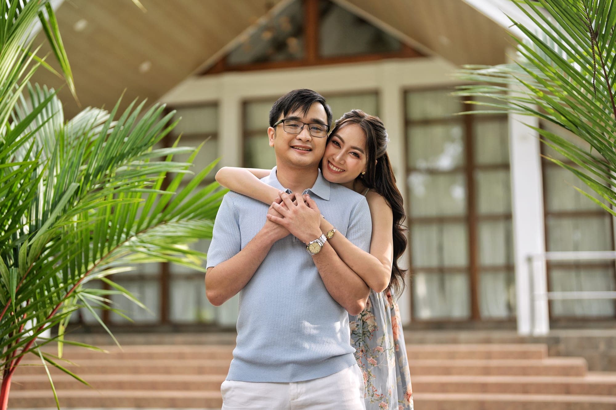 Justin and Lovely embracing and smiling at the camera in front of a timber-roofed pavilion, Lovely resting her chin on his shoulder