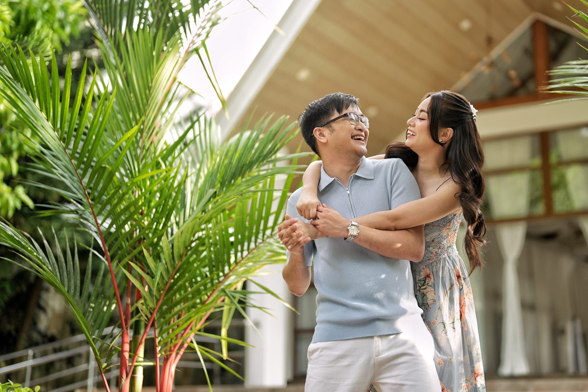 Lovely laughing with her arms around Justin from behind, framed by tropical palm leaves in front of a timber-roofed pavilion