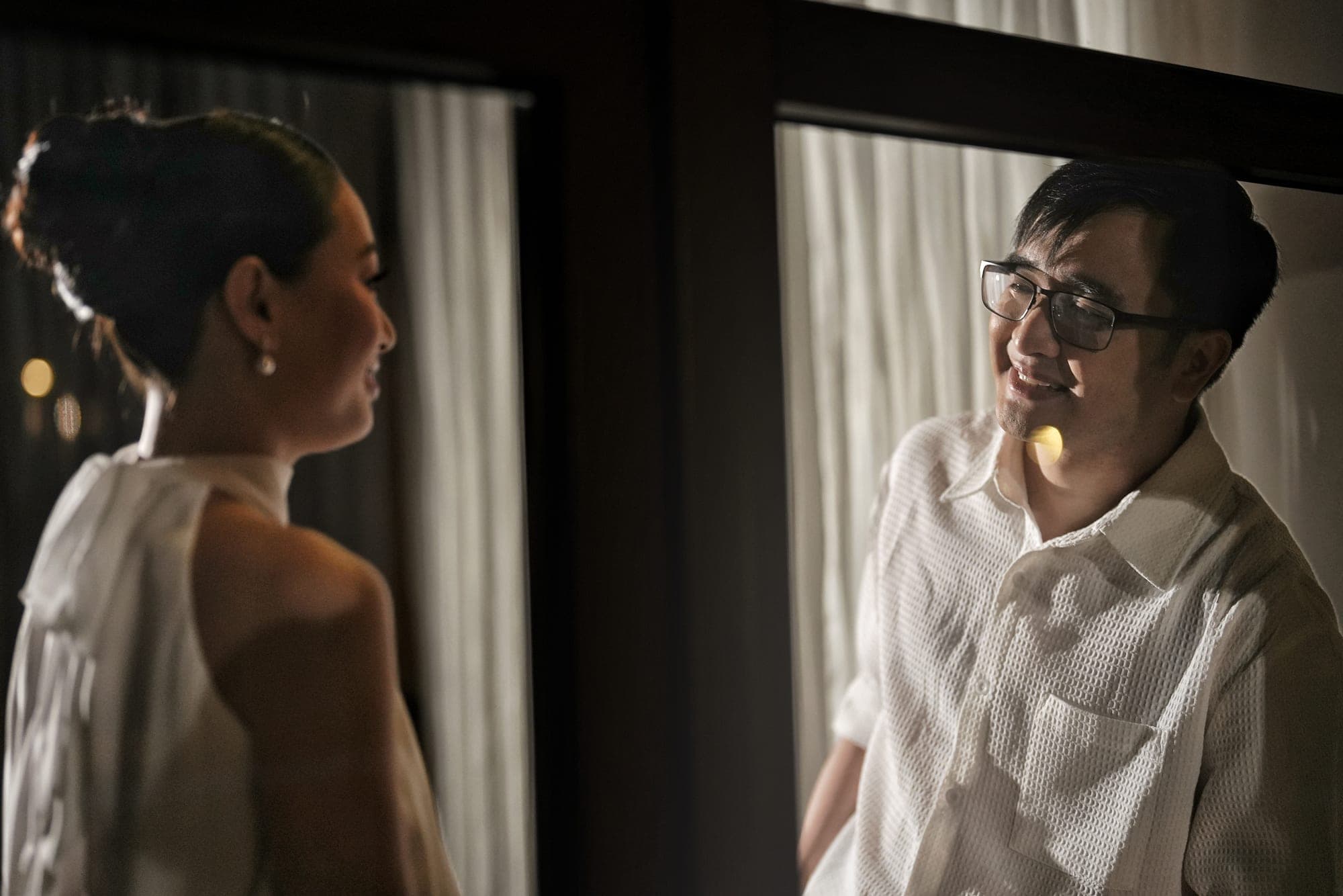 Justin, in a light shirt, and Lovely, in a bare-shoulder gown, smiling at each other in a dim room reflected in a window pane