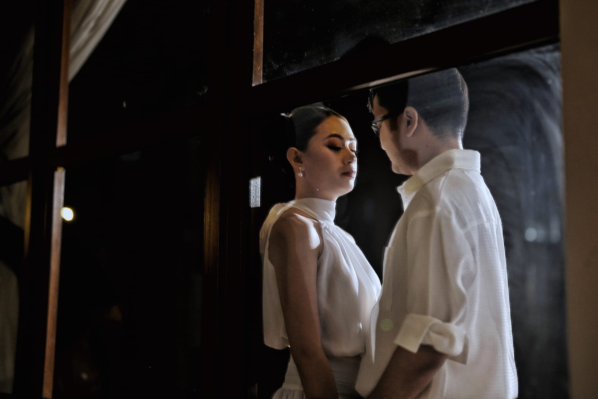Justin and Lovely standing close together in dramatic low light, foreheads nearly touching, both dressed in white