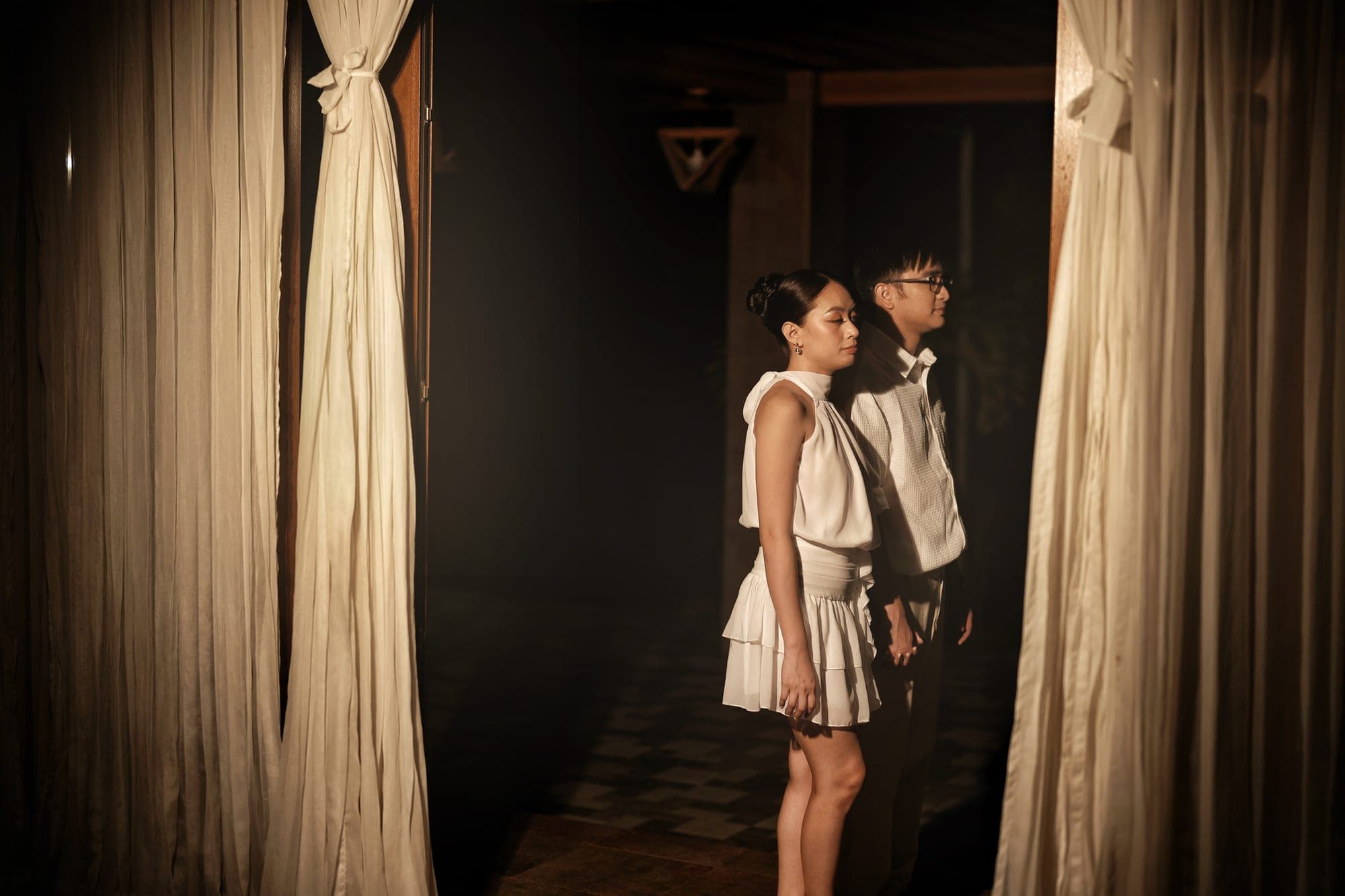 Justin and Lovely standing side by side in a candlelit doorway framed by draped curtains, both dressed in white