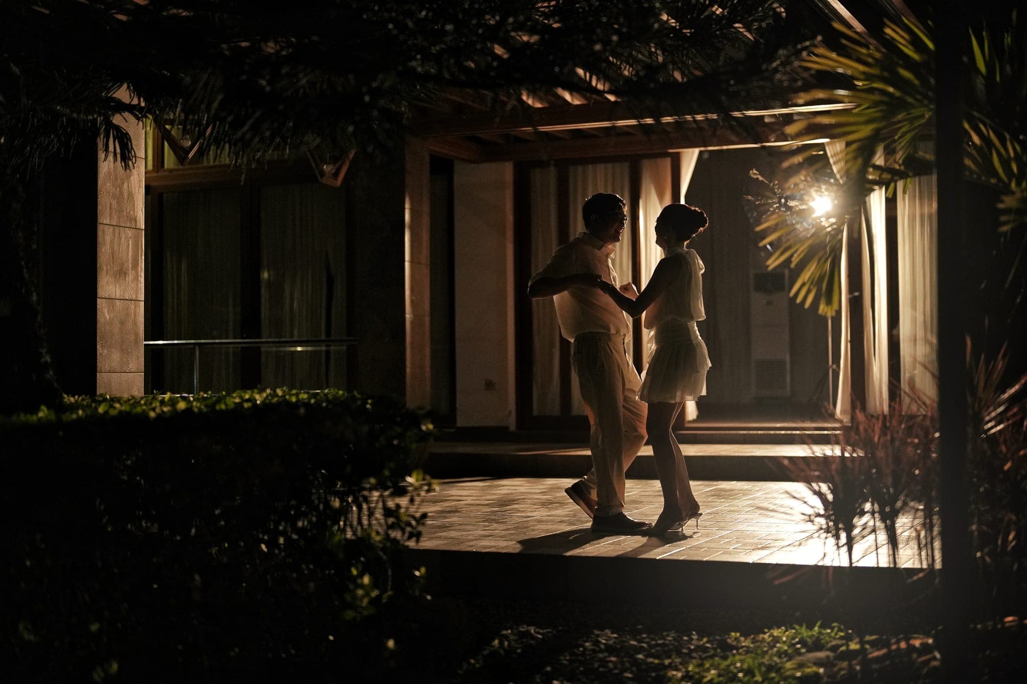 Justin and Lovely facing each other on a lantern-lit patio at night, silhouetted against warm light spilling from the building behind them