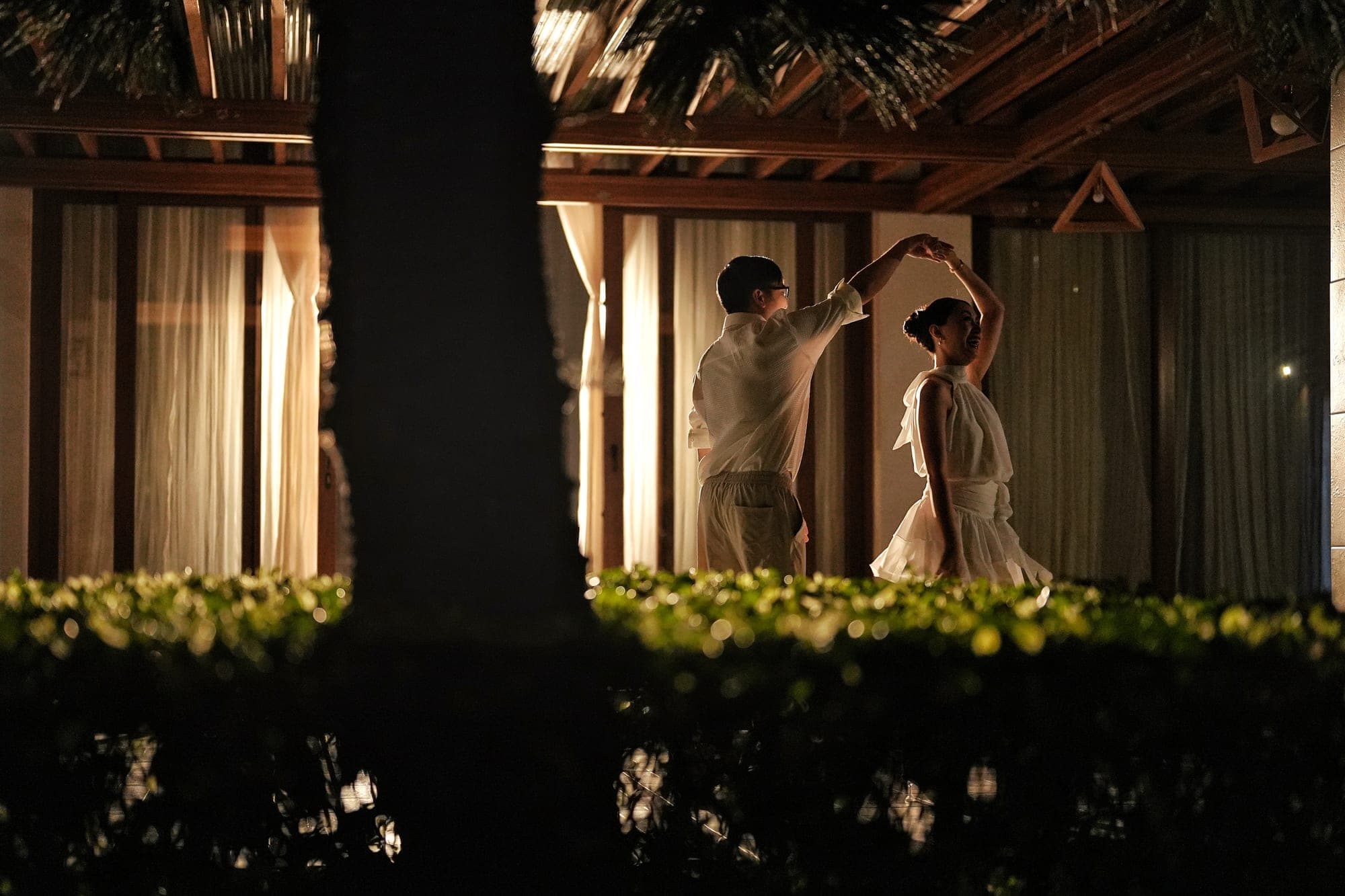 Justin twirling Lovely under a lit pavilion at night, her dress flaring out mid-spin, framed by a dark hedge in the foreground
