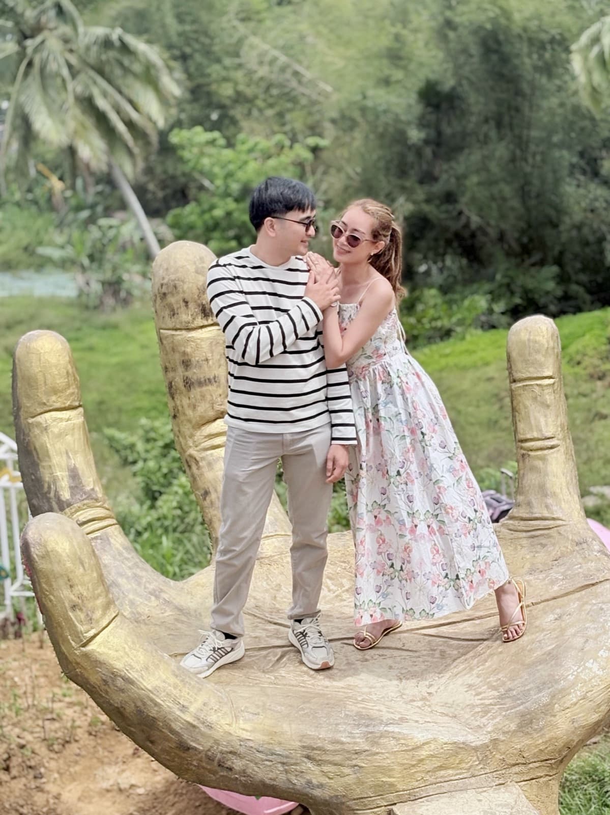 Justin and Lovely embracing atop a giant carved golden hand sculpture, surrounded by palm trees and greenery